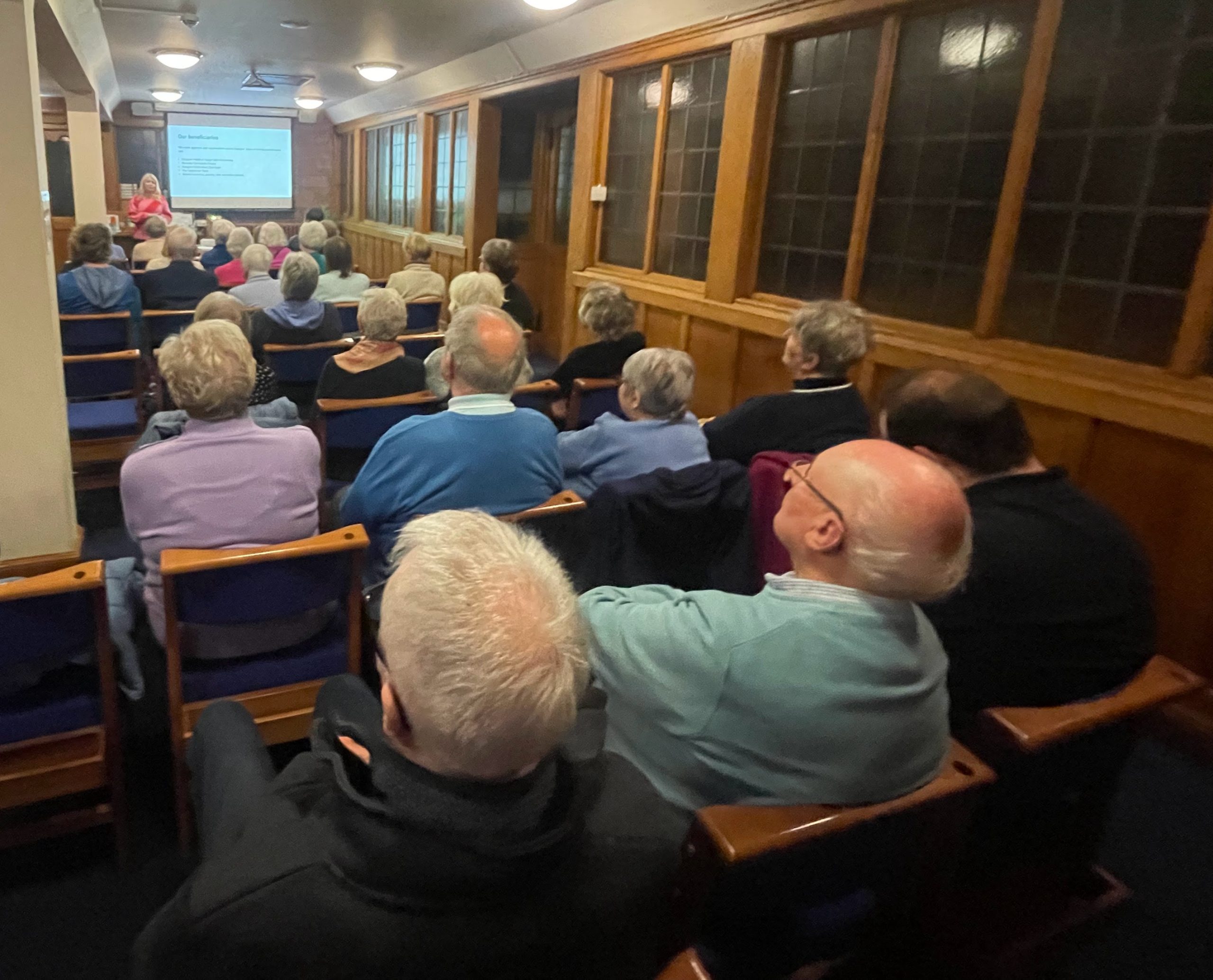 People sitting in rows of blue chairs surrounded by wood panelled walls watching a powerpoint presentation
